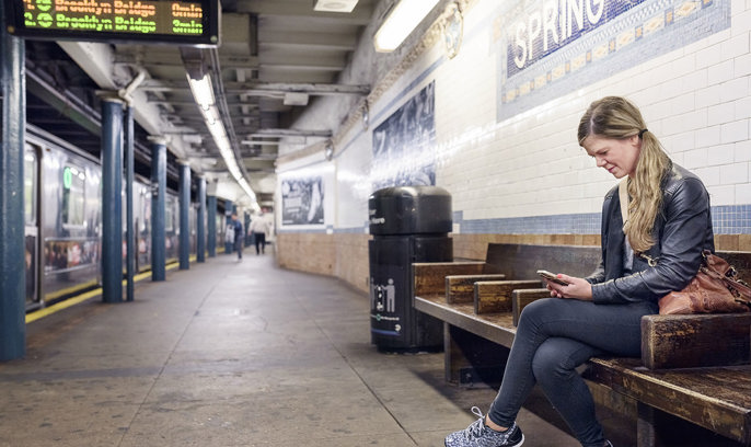 En ung kvinna sitter på en t-banestation i NY och tittar på sin mobil.