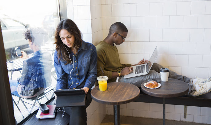 Två ungdomar sitter och tittar på surfplatta och laptop på ett café